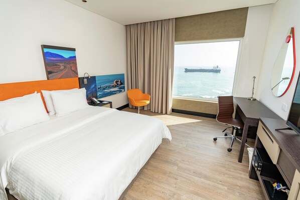 Habitación de hotel con cama, escritorio, silla, televisor y ventana grande con vista al mar.