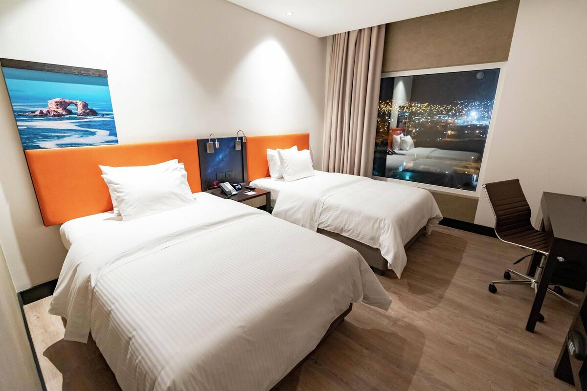 Une chambre d’hôtel avec deux lits, un bureau, une chaise, une table de chevet et une grande fenêtre donnant sur la ville.