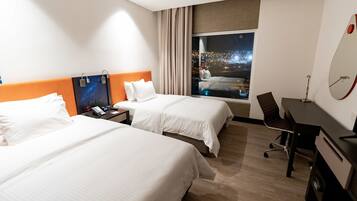 A hotel room with two beds, a desk, a chair, a nightstand, and a large window with a city view.