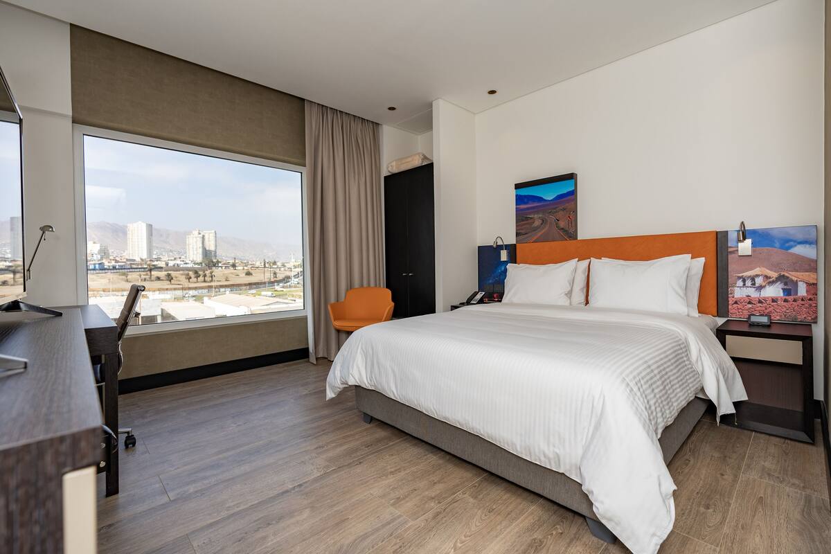 Een hotelkamer met een groot bed, een bureau, een stoel en uitzicht op een stadsgezicht door grote ramen.