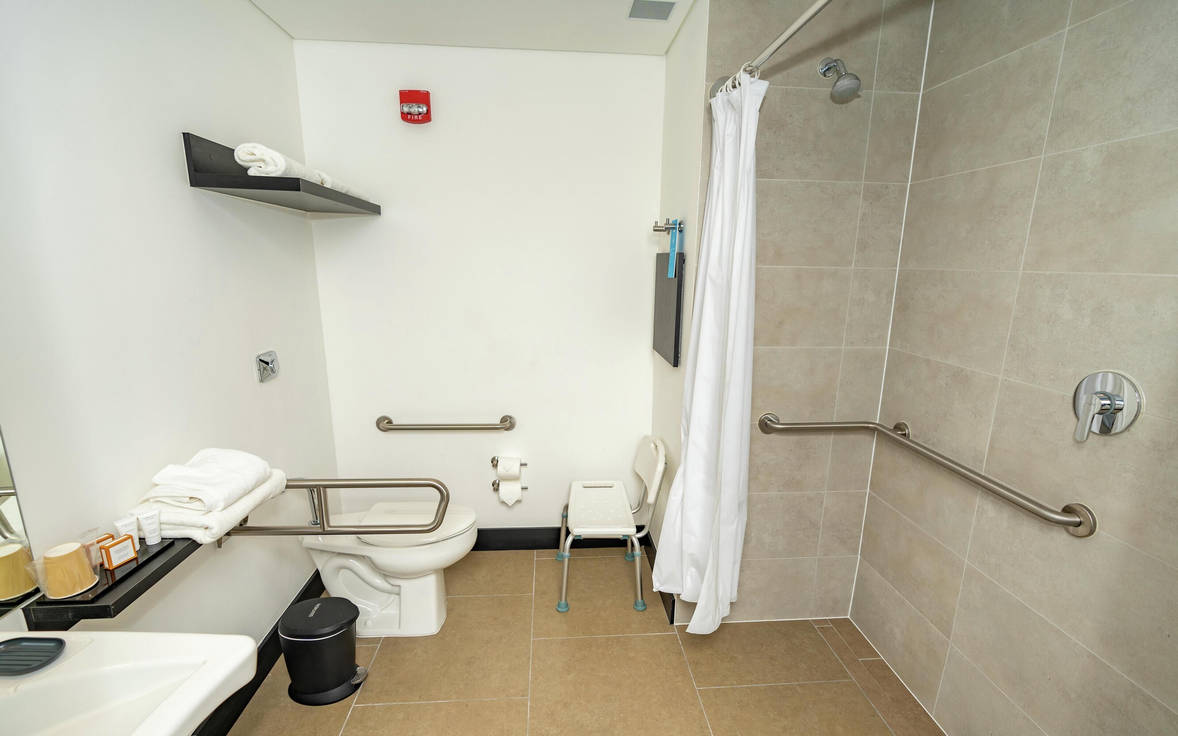 A bathroom with a toilet, shower, grab bars, a trash bin, and a towel rack.