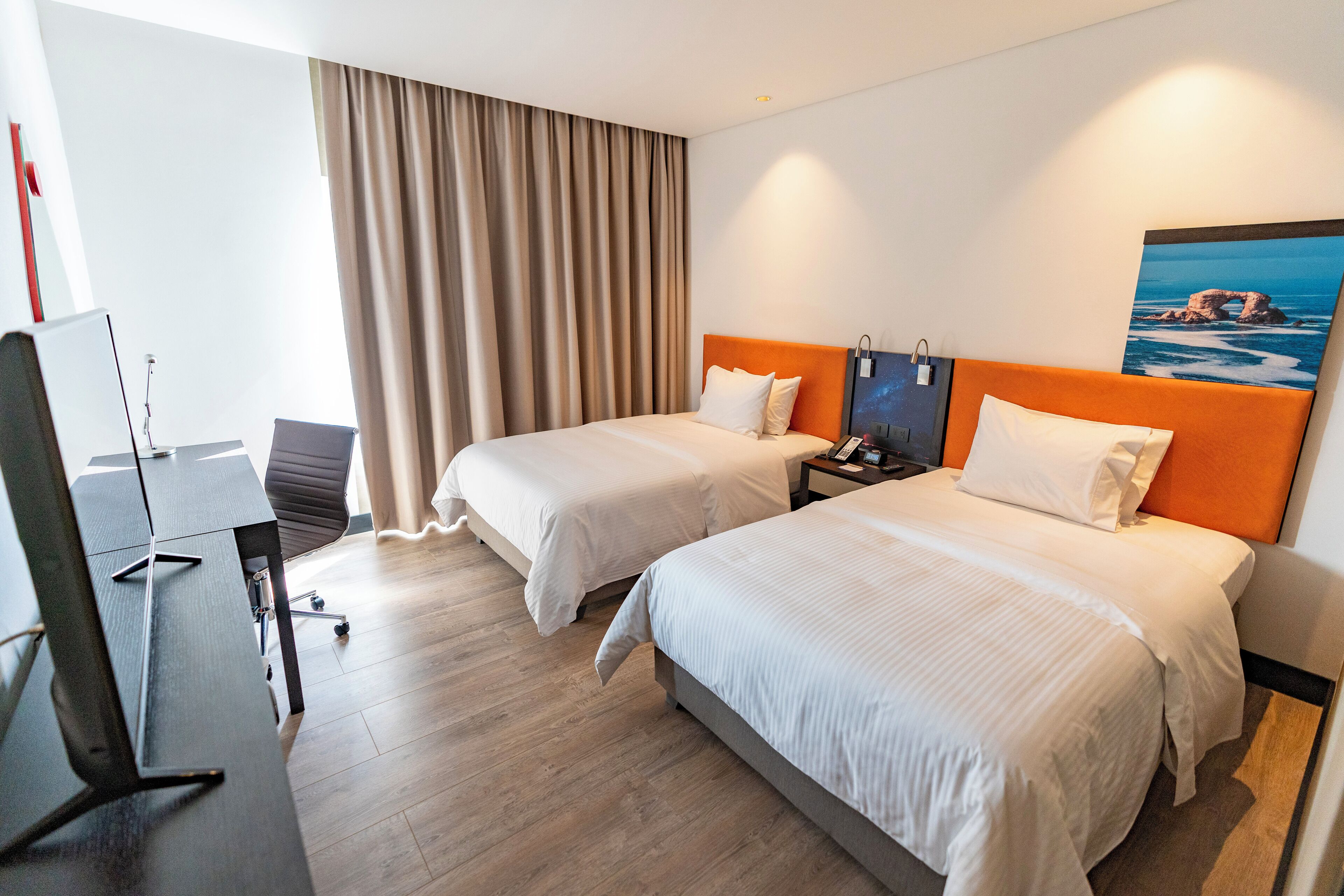 Quarto de hotel com duas camas, uma escrivaninha, uma cadeira, uma televisão e um quadro na parede.