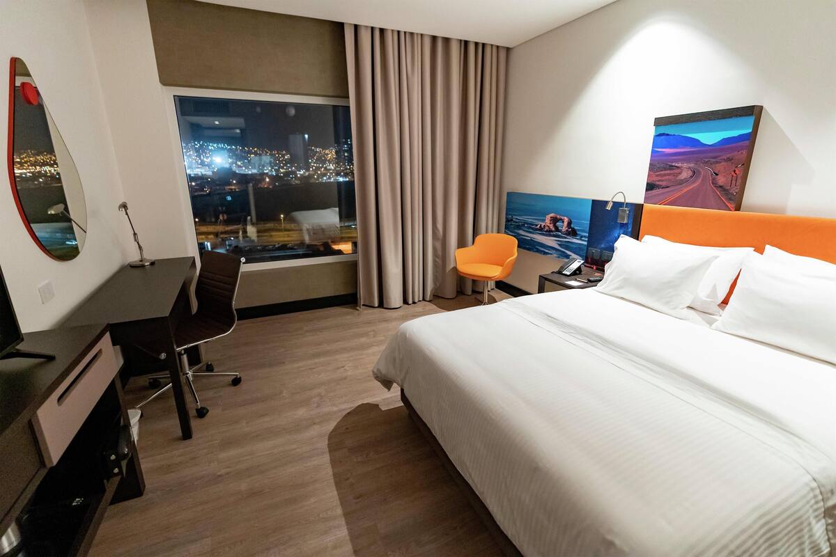Hotelkamer met een bed, een bureau, een stoel, een groot raam met uitzicht op de stad en een schilderij aan de muur.