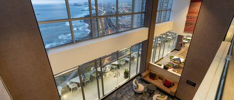 Habitación de hotel en un edificio alto con un amplio ventanal con vista al océano, un separador de vidrio que divide la habitación del área de comedor inferior, y una decoración interior moderna.
