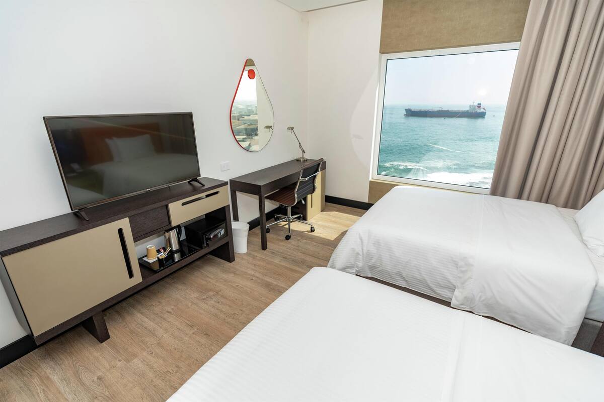 Een hotelkamer met een bed, een bureau met een televisie, een spiegel en uitzicht op zee.
