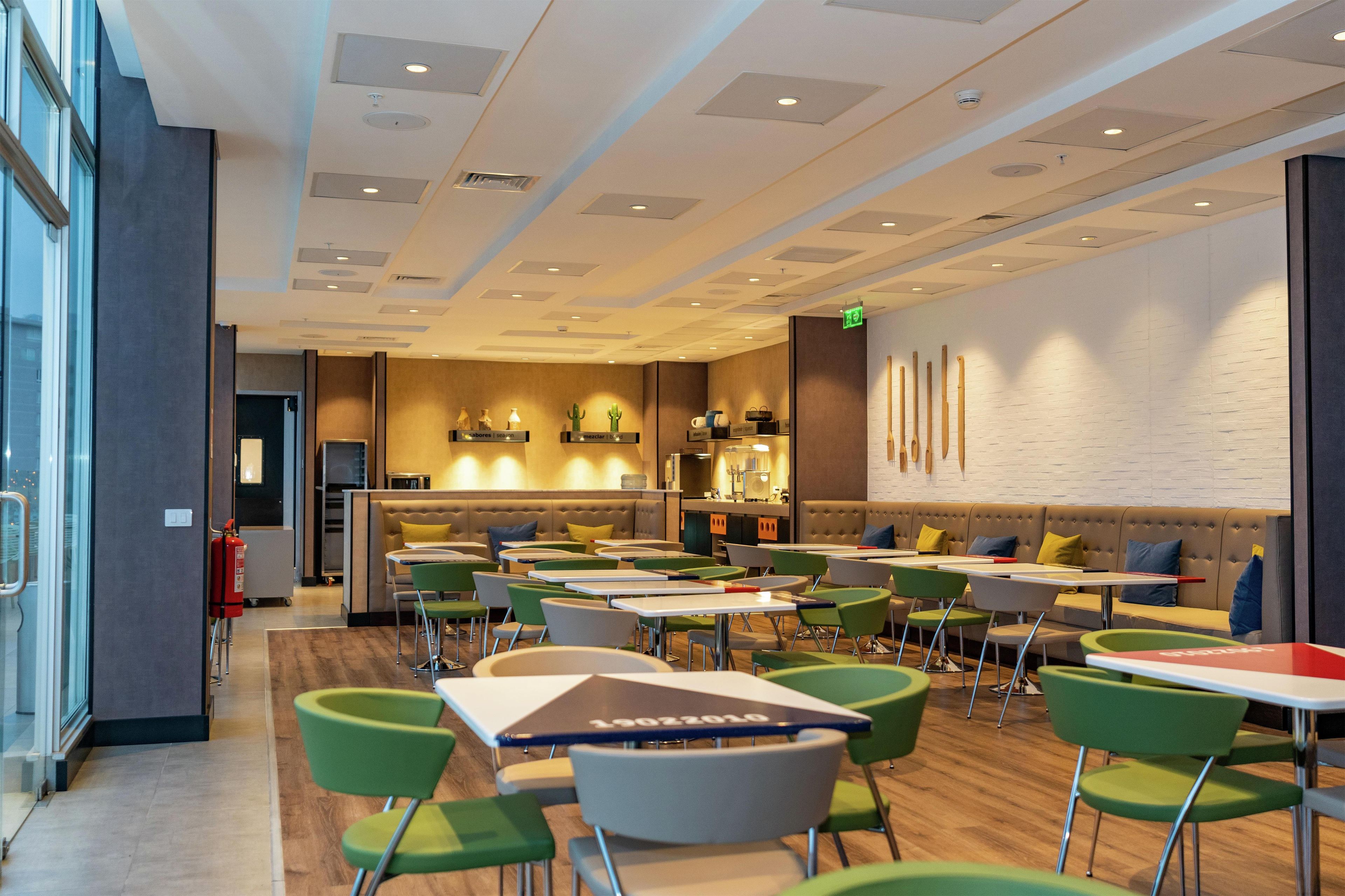 A modern dining area with tables and chairs, a kitchen in the background, and a brick wall with a light fixture.