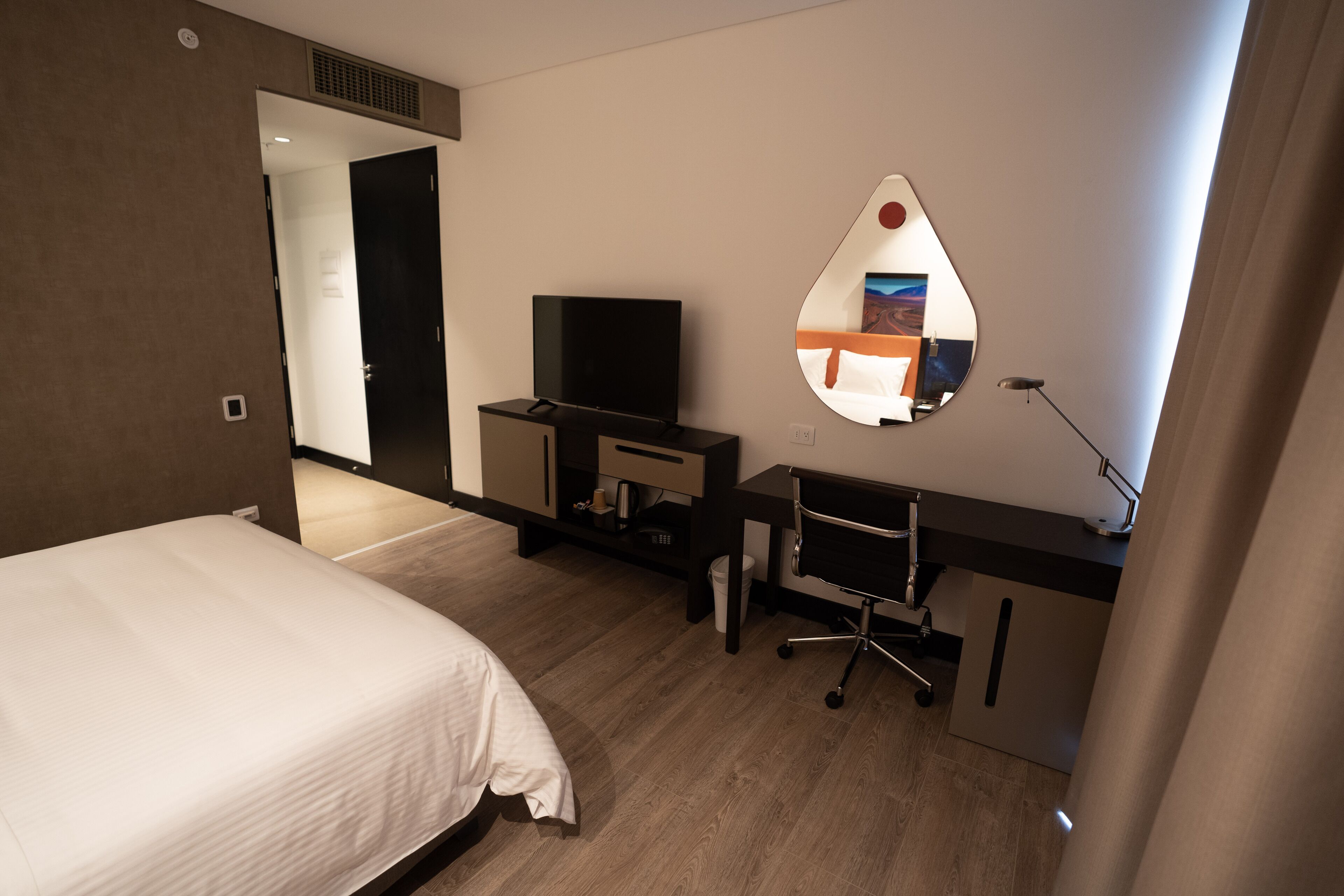 Una habitación de hotel con cama, escritorio, silla, televisor y espejo.