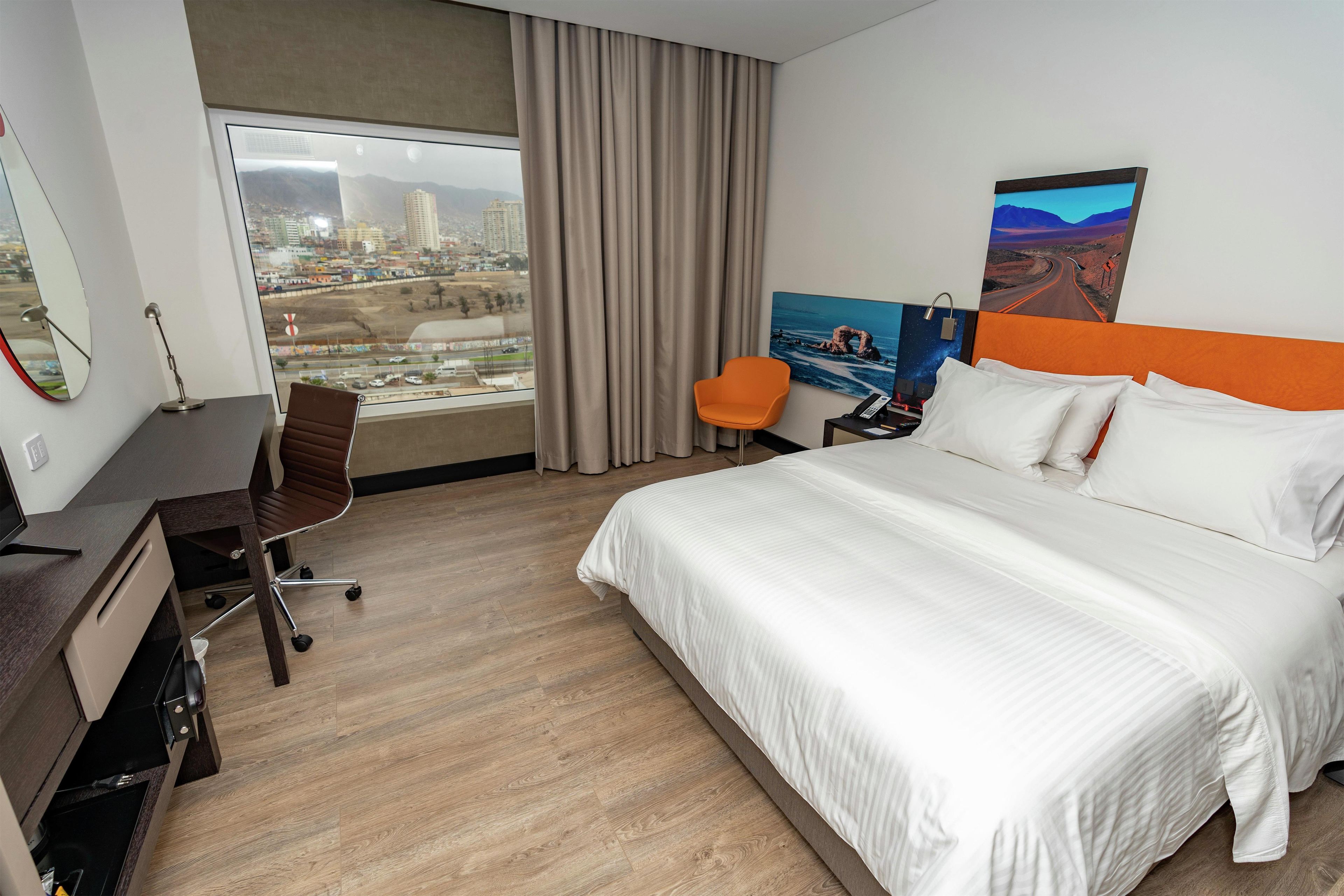 A hotel room with a large bed, a desk, a chair, and a view of a cityscape through a window.