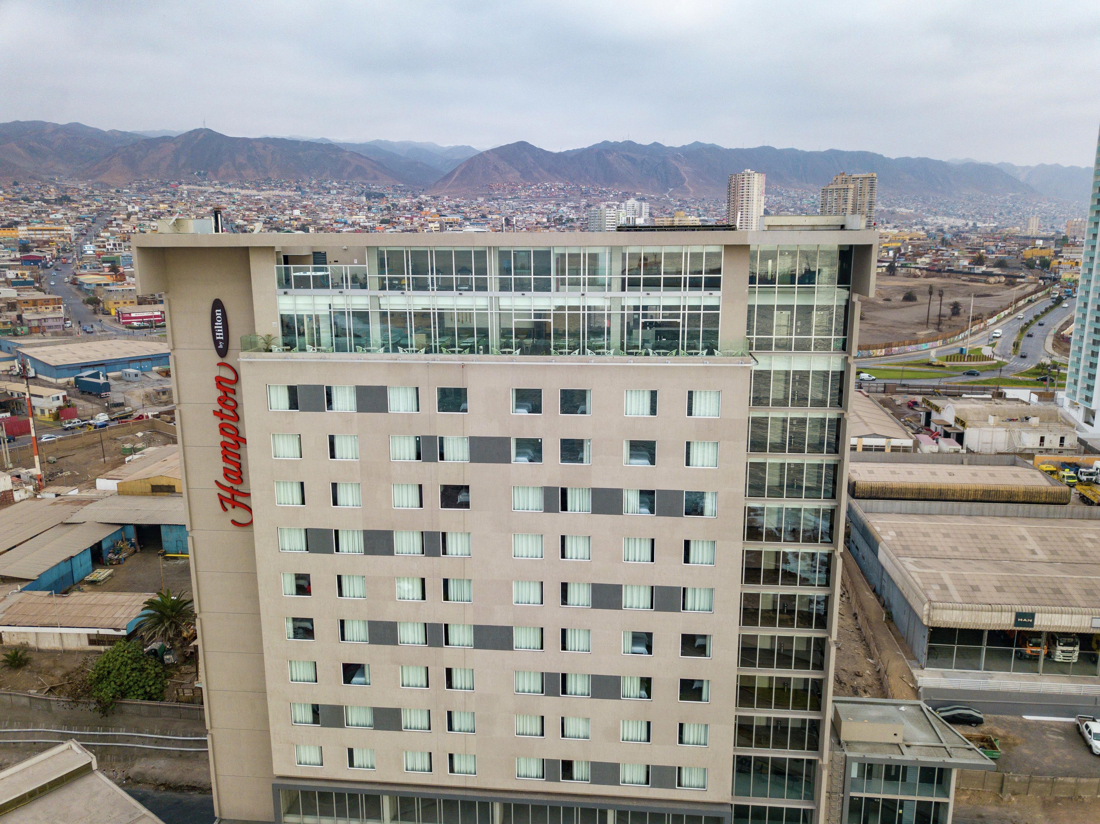 Un hotel de varios pisos con un llamativo letrero de Hilton, amplias ventanas y vistas a un paisaje urbano y montañas al fondo.