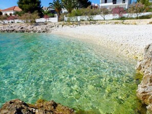 A clear coastal beach with a rocky shoreline and a white building in the background. - Apartments Srđan, (15477), Zecevo, hr (Zecevo)