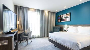 A modern hotel room with a large bed, a desk, a chair, and a TV.