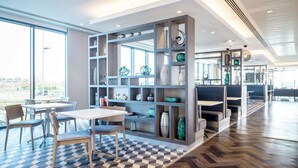 A modern interior with a checkered floor, a central seating area with a table and chairs, and a decorative bookshelf with various items. - Hampton by Hilton Stockton on Tees (Stockton-on-Tees)