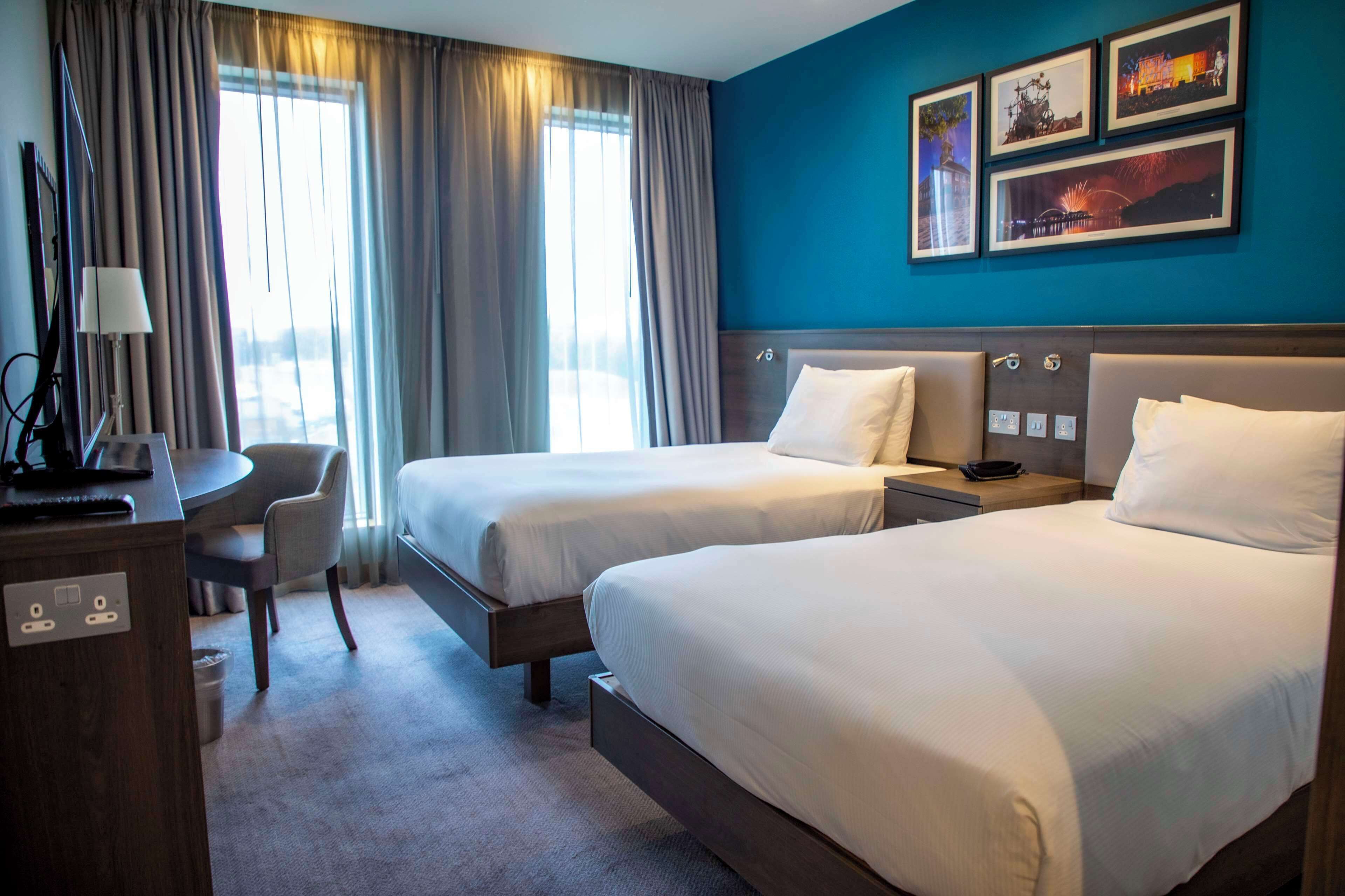 A hotel room with two beds, a desk, a chair, and a view of the outdoors through large windows.