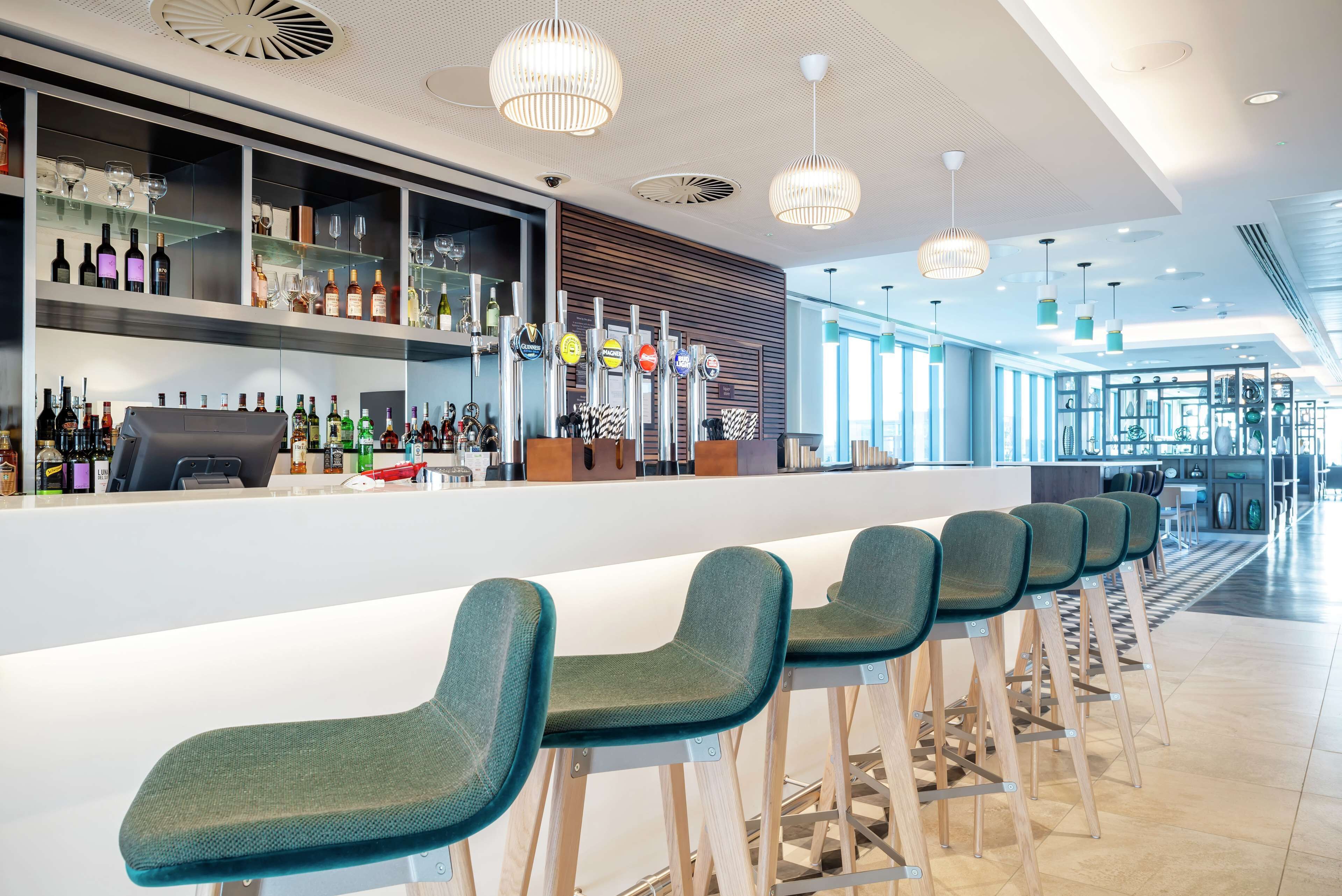 Eine moderne Bar mit einer langen Theke, mehreren Hockstühlen und einer Auswahl an Flaschen und Gläsern auf Regalen.