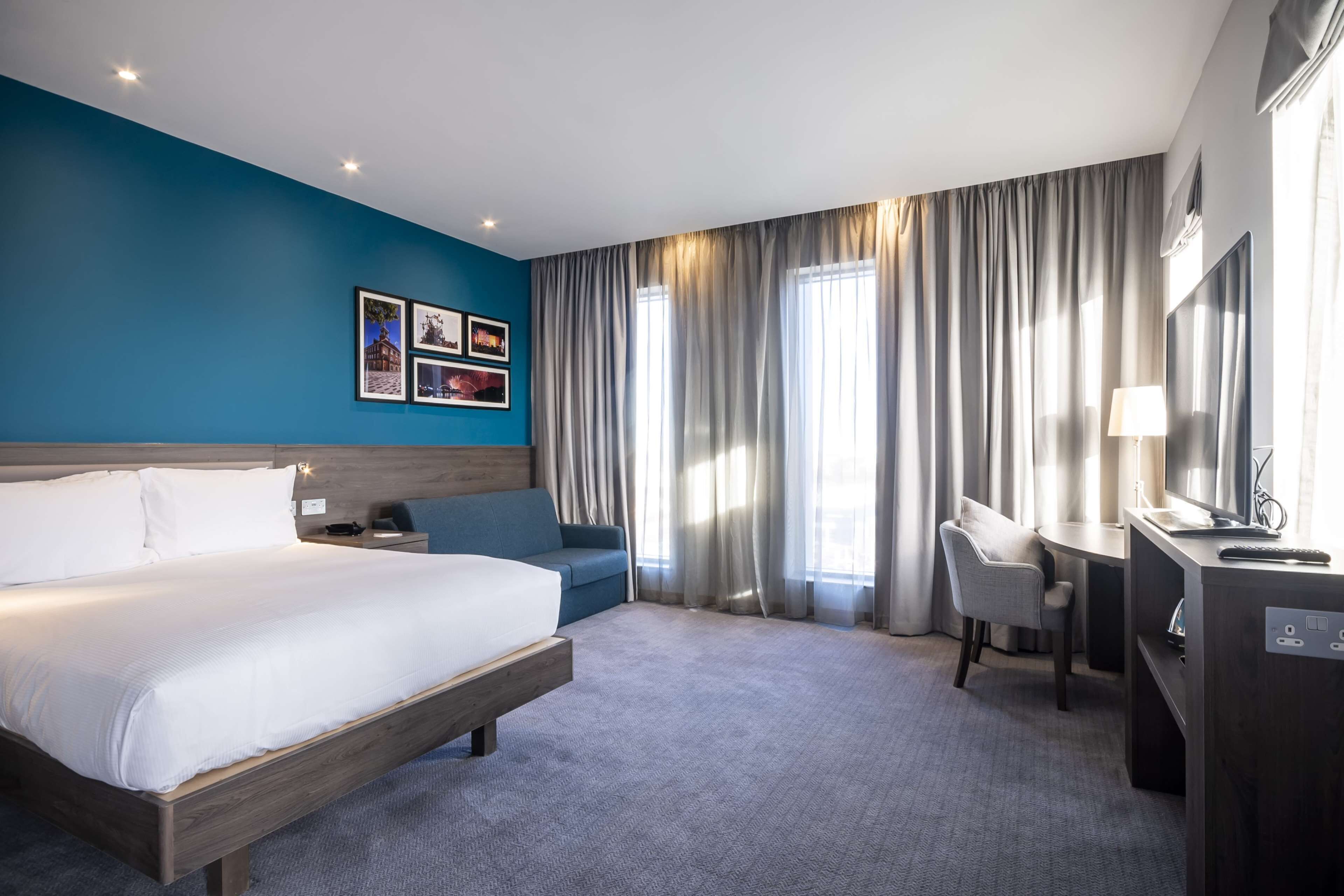 A hotel room with a large bed, a desk with a chair, a TV, and a desk lamp.