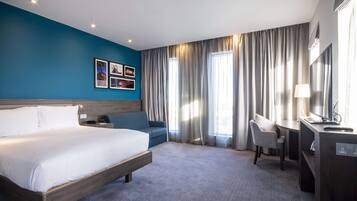 A hotel room with a large bed, a desk with a chair, a TV, and a desk lamp.