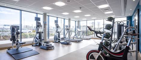 Un gimnasio moderno con máquinas de cardio, pesas y ventanas amplias con vista a la ciudad.