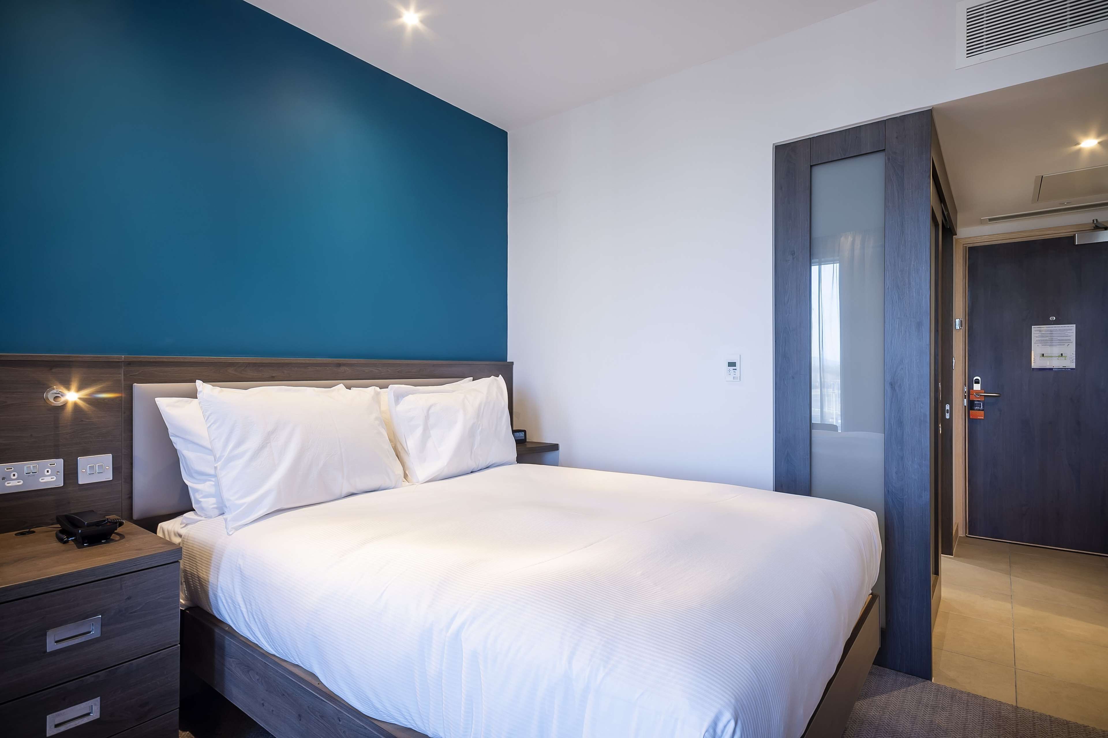 A modern hotel room with a large bed, a wooden nightstand, and a blue accent wall.
