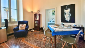 Un comedor con una mesa preparada para dos personas, un sillón azul, una estantería y un amplio ventanal con vista a palmeras.