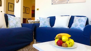 Una sala de estar con un sofá azul, una mesa de centro con fruta y un cuadro con motivos playeros.