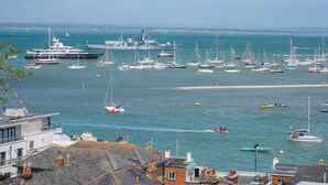 Marina com vários barcos e iates atracados, praia arenosa ao fundo e edifícios com telhados visíveis.