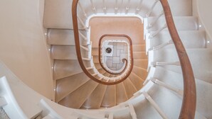 Escadaria em espiral com corrimão de madeira e balaústres brancos, que leva a um patamar com um pequeno objeto decorativo.