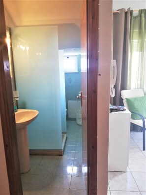 Une salle de bain avec un lavabo, des toilettes et un espace douche. On y trouve une chaise et une petite table avec un téléphone et un livre.