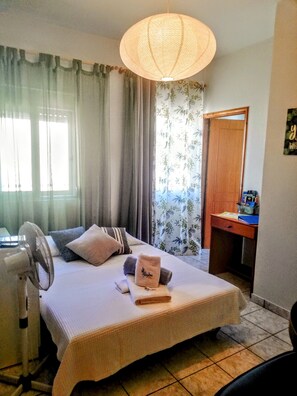 Une chambre à coucher avec un lit, un bureau, un ventilateur sur pied et une fenêtre avec des rideaux.