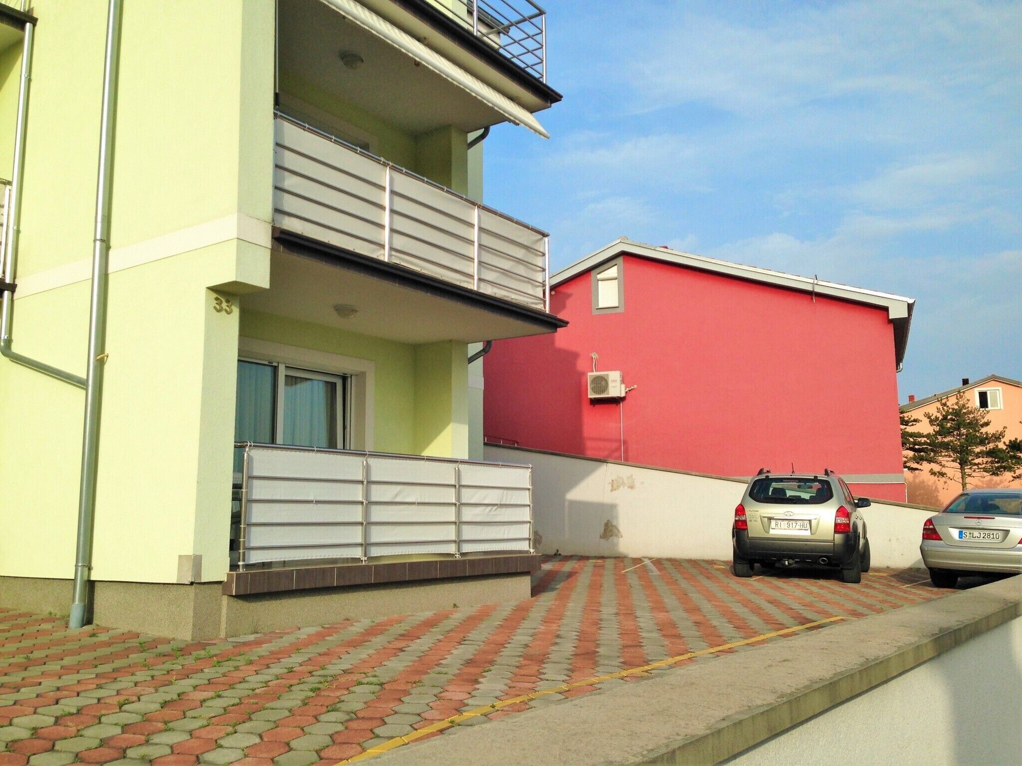 Un immeuble résidentiel doté d’un balcon, avec une voiture garée devant et un mur rouge.