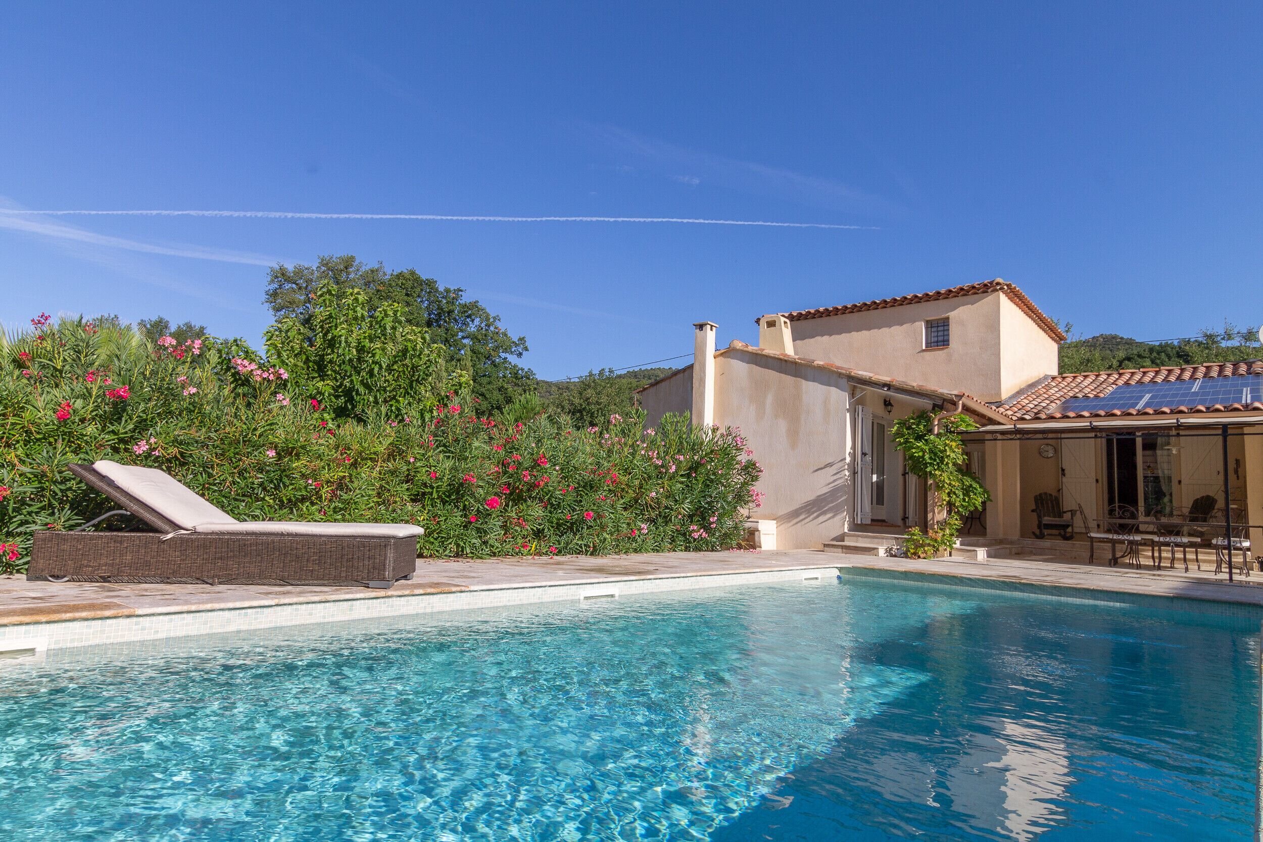 A clear blue swimming pool with a lounge chair, a house with a tiled roof, and lush greenery in the background.