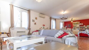 A living room with a sofa, ottoman, and a glass coffee table. There is a bookshelf and a dining area with chairs. - Macs Reve - A dream for bikers : 2 bikes for men & 2 bikes for women available for free. (Collobrières)