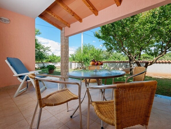 A covered patio with a round table and wicker chairs, a lounge chair, and a fruit basket.