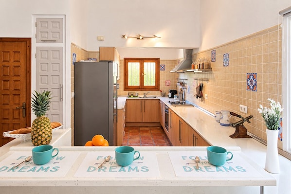 A kitchen with wooden cabinets, a refrigerator, a window, and a countertop with a pineapple and oranges.