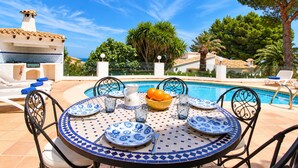 Une table ronde, ornée de vaisselle aux motifs bleus et blancs, un saladier rempli d’oranges et, en arrière-plan, une piscine d’un bleu limpide.