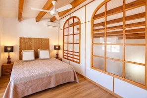 A bedroom with a bed, two bedside tables, a ceiling fan, and large windows with wooden frames.