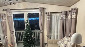 Een gezellige woonkamer met een bank, voetenbankje en een klein bijzettafeltje. Achterin staat een kerstboom en er is een raam met gordijnen.