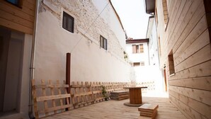 A narrow wooden corridor with a rustic stone wall on one side and a wooden fence on the other. A round wooden table and a stack of wooden planks are placed on the floor.
