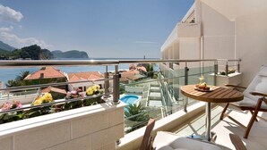 A balcony with a glass railing, a wooden table with a plate of fruit, and a view of the sea and buildings in the background. - Monte Casa Spa And Wellness (Petrovac)