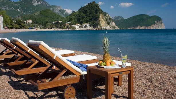 A beach with wooden lounge chairs and a table with fruit and a drink, overlooking a coastal area with mountains in the background. - Monte Casa Spa And Wellness (Petrovac)