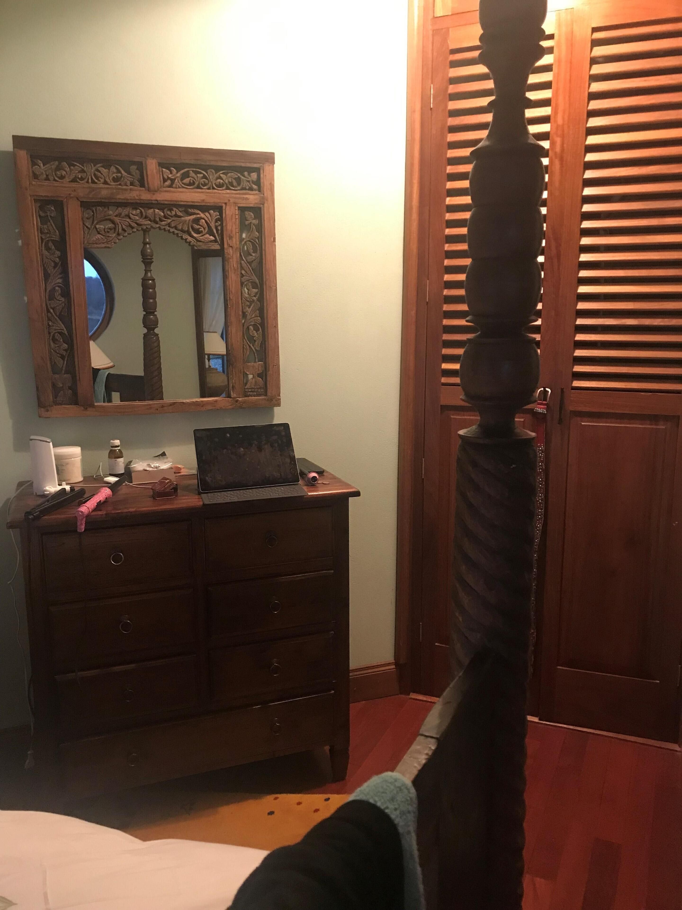 Une chambre à coucher avec une commode en bois, un miroir orné de sculptures, un ordinateur portable et une porte en bois agrémentée d’un motif décoratif.