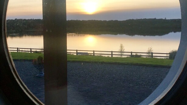 A view through a circular window of a sunset over a calm lake, with a wooden fence and grassy area in the foreground. - Idyllic and Unique House on the Lake  (Whitegate)