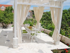 Relax in a serene outdoor space with a pergola, tables, and chairs, surrounded by lush greenery and a residential view.