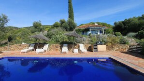 A swimming pool with lounge chairs, a stone wall, and a house with a tiled roof in the background.