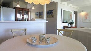 A dining area with a round table, chairs, and a tray with decorative cups. There is a kitchen area with cabinets and a refrigerator in the background.