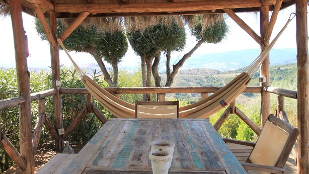 A wooden deck with a hammock and a table, overlooking a mountainous landscape.