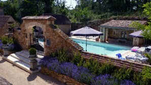 Una piscina de piedra rodeada de sillones de descanso y sombrillas, con edificios de piedra y un muro de piedra al frente.