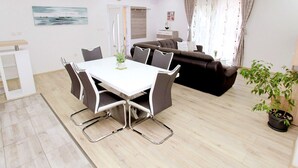 Ein modernes Wohnzimmer mit Essbereich, ausgestattet mit einem weißen Tisch und Stühlen, einem braunen Sofa und einer Topfpflanze.