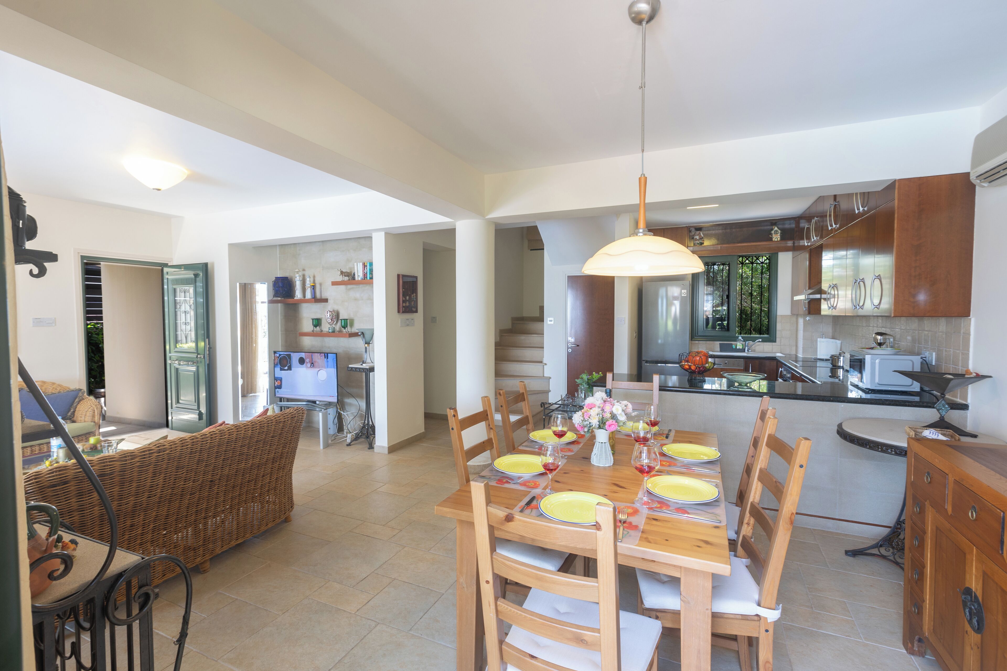 Uma ampla sala de jantar com uma mesa de madeira posta para seis pessoas, cercada por cadeiras. Uma cozinha com eletrodomésticos modernos é visível ao fundo.
