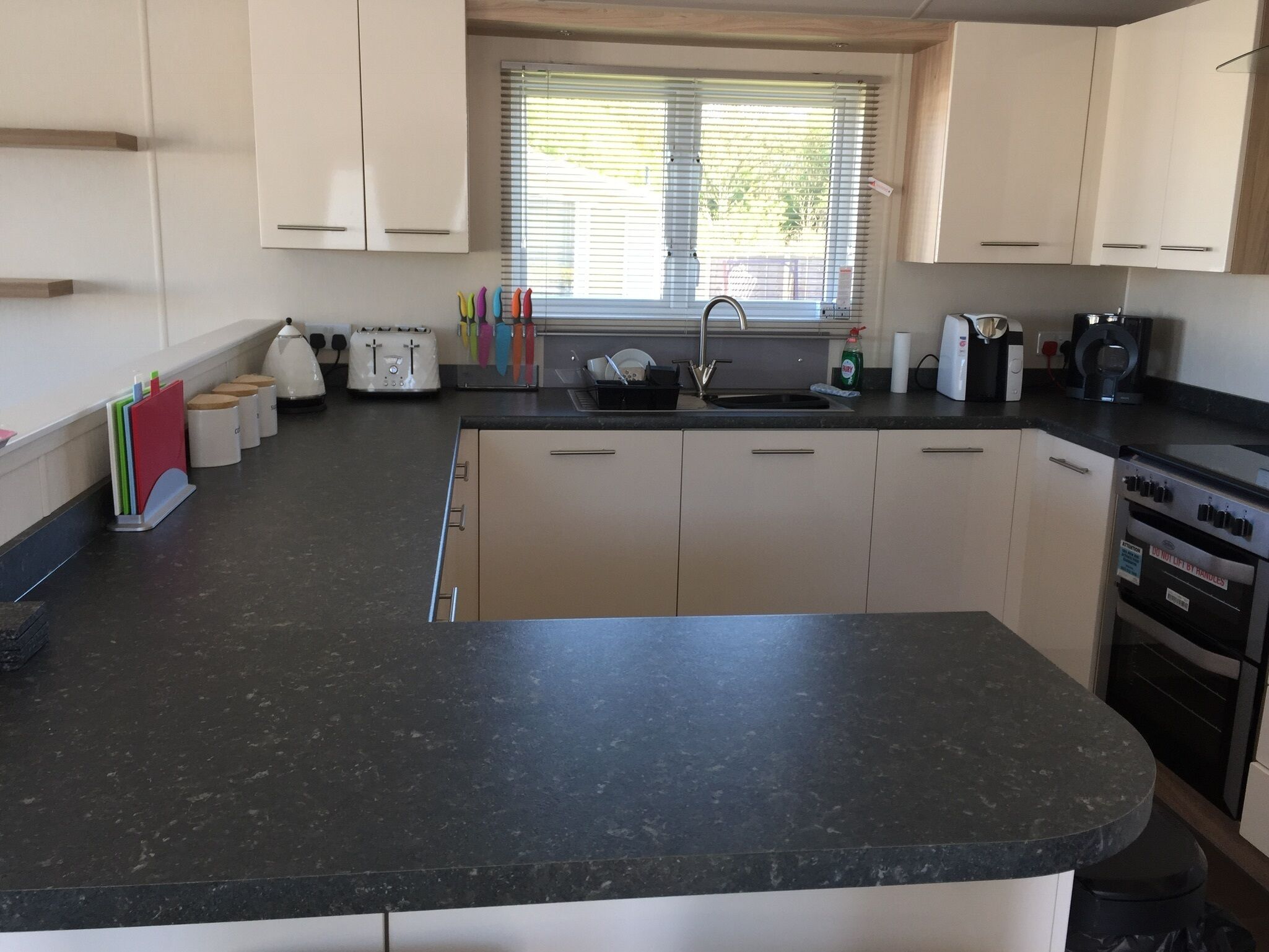 A modern kitchen with a black countertop, white cabinets, a window with blinds, a sink, a stove, and various kitchen appliances.