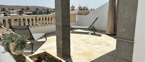 A covered outdoor patio with stone flooring, a seating area with a chair, and planters with plants.